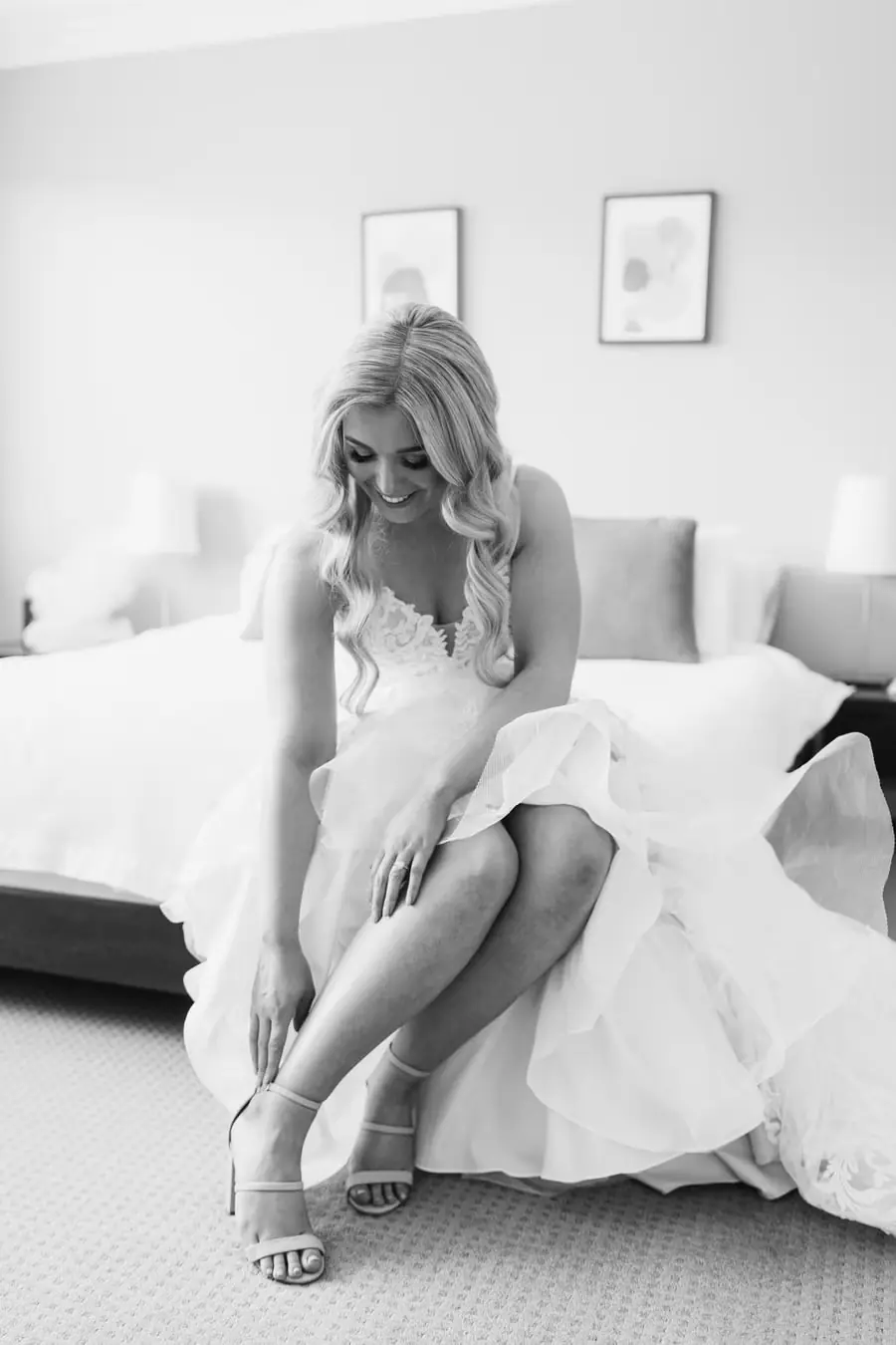 Black-and-white photograph of the bride fastening a shoe while seated on a bed