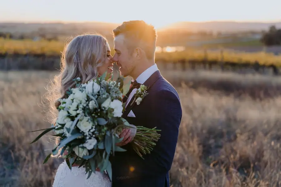 Bride and groom kissing behind a bouquet in a sunlit paddock