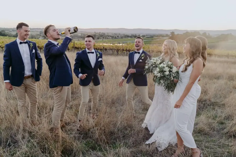 Groomsman raising a bottle to his mouth as the wedding party laughs
