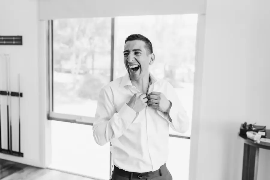 Black and white photograph of the groom laughing while fastening his white shirt