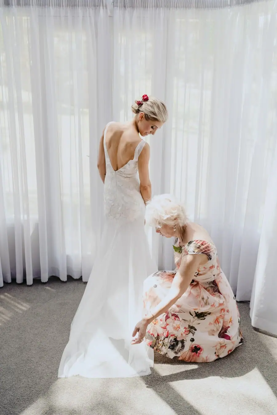 Woman kneeling to adjust the bride's dress beside a curtained window