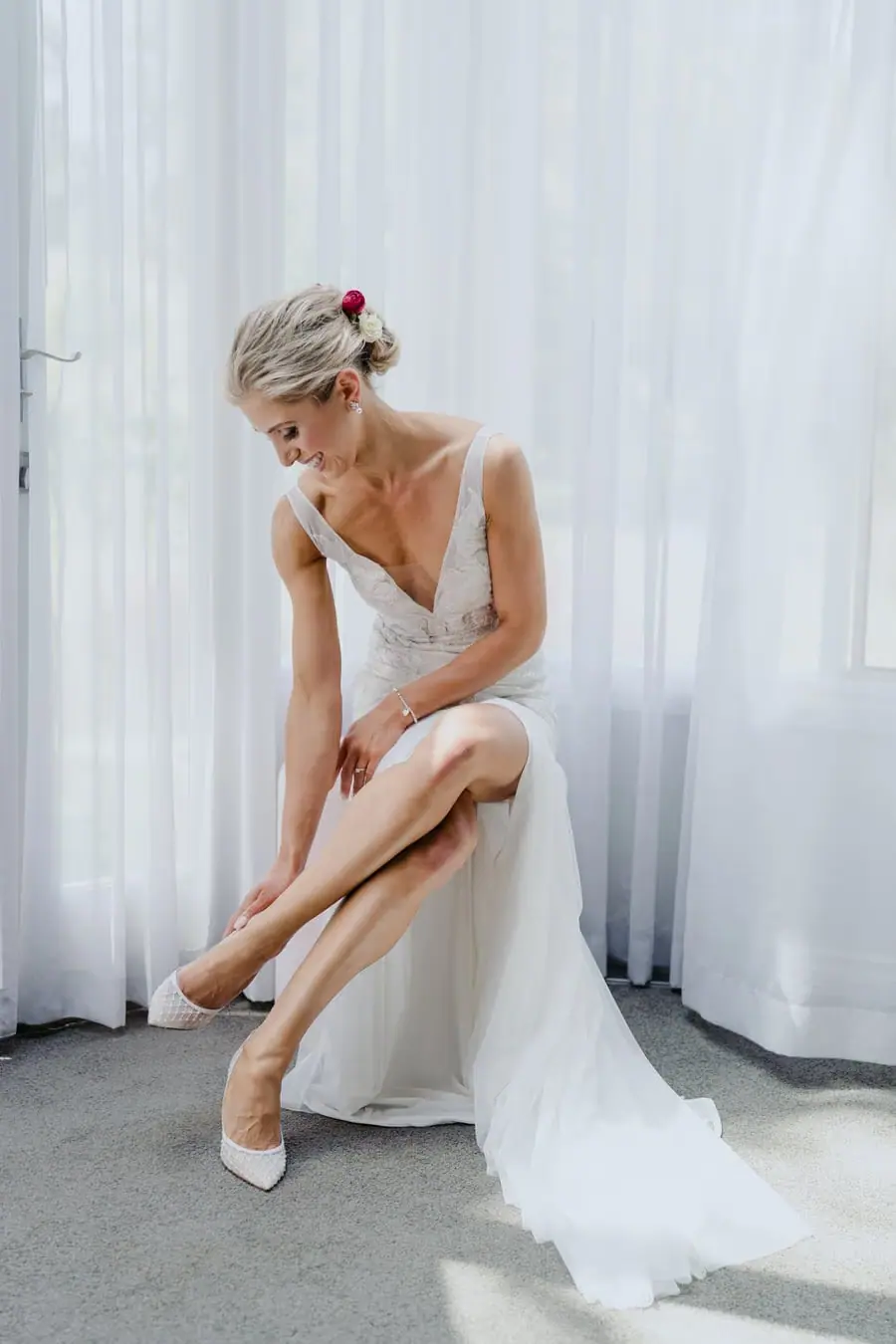 Bride fastening a white shoe while seated beside a window