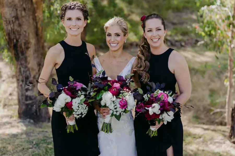 Bride and two bridesmaids in black dresses holding colourful bouquets beneath trees
