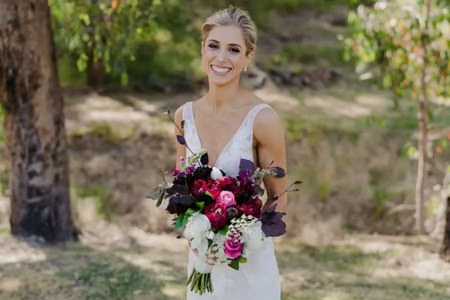 Bride smiling with a burgundy and white bouquet beneath a tree