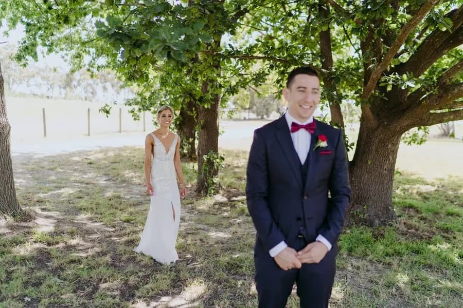 Bride approaching the groom from behind beneath a leafy tree