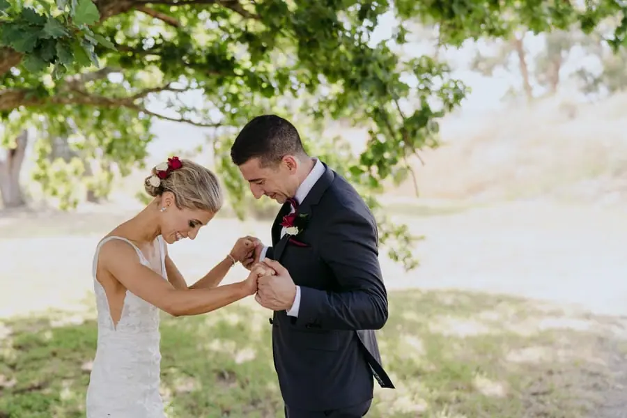 Bride and groom smiling down while holding hands beneath a tree