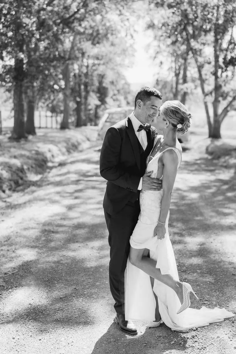 Black and white photograph of the groom kissing the bride's forehead on a gravel road