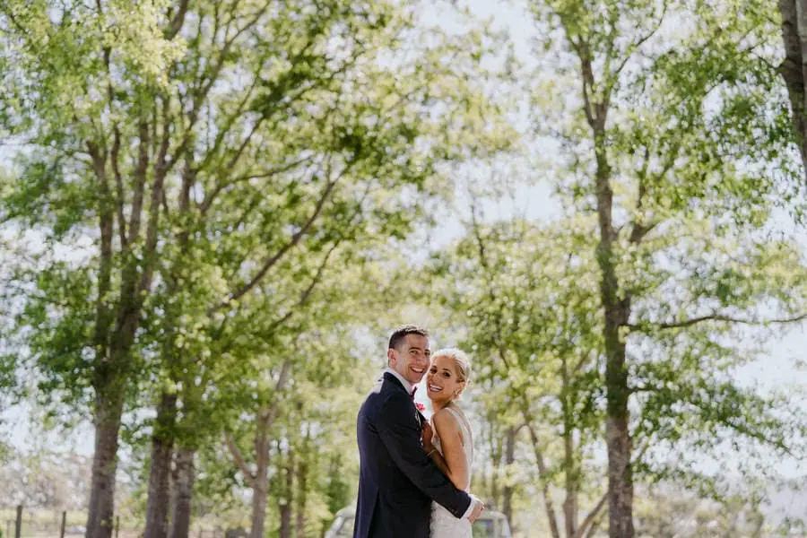 Bride and groom embracing beneath tall leafy trees
