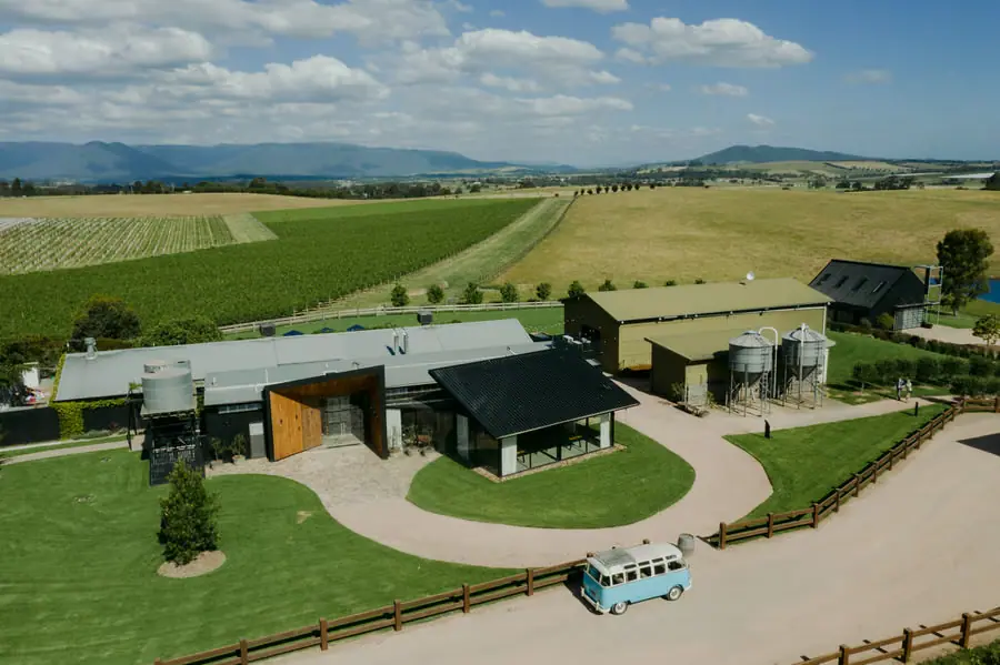 Aerial view of a dark-roofed building and curved path surrounded by farmland and vineyard rows
