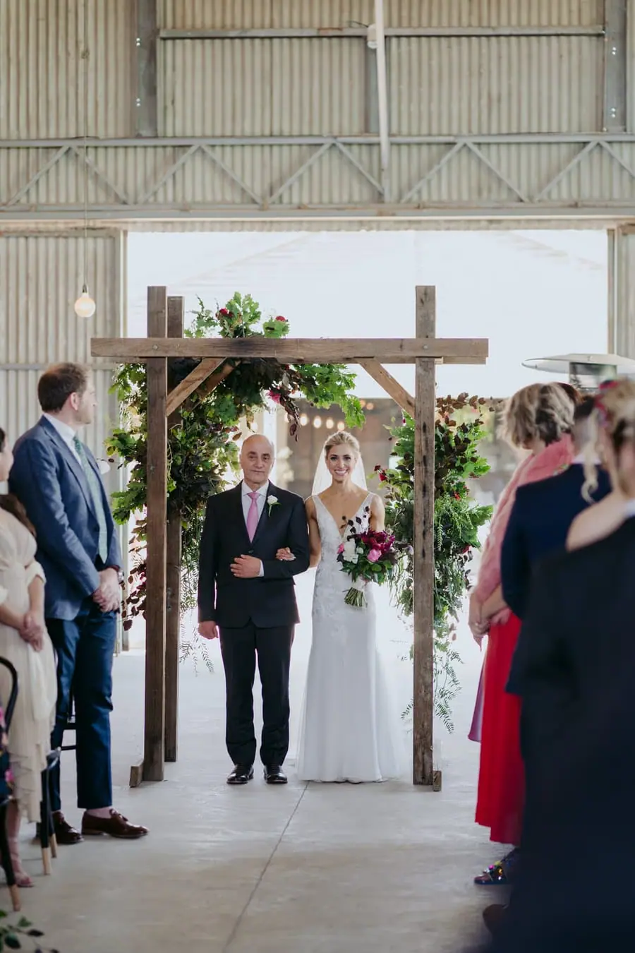 Bride walking into an indoor ceremony beside a man beneath a timber floral arch