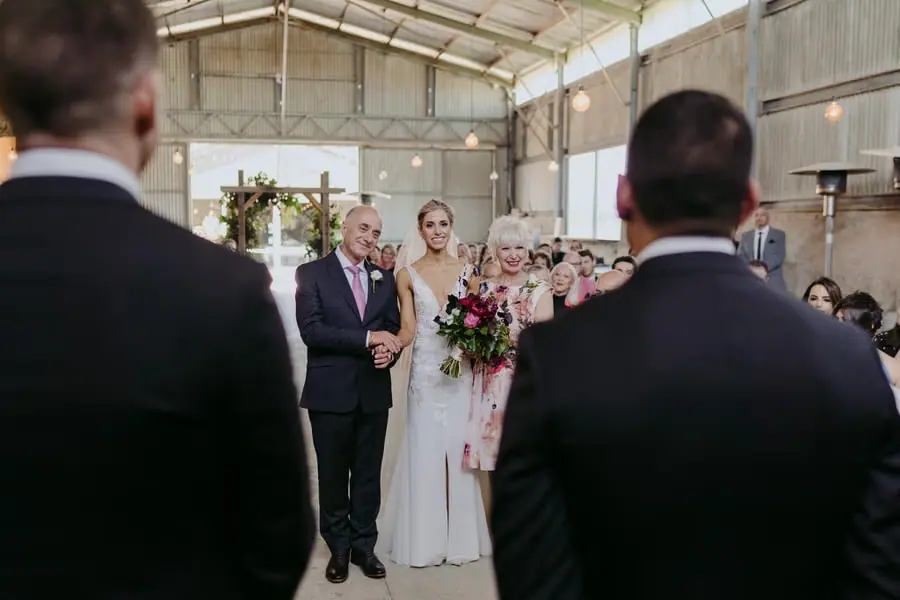 Bride entering the ceremony with a man and woman as the groom watches