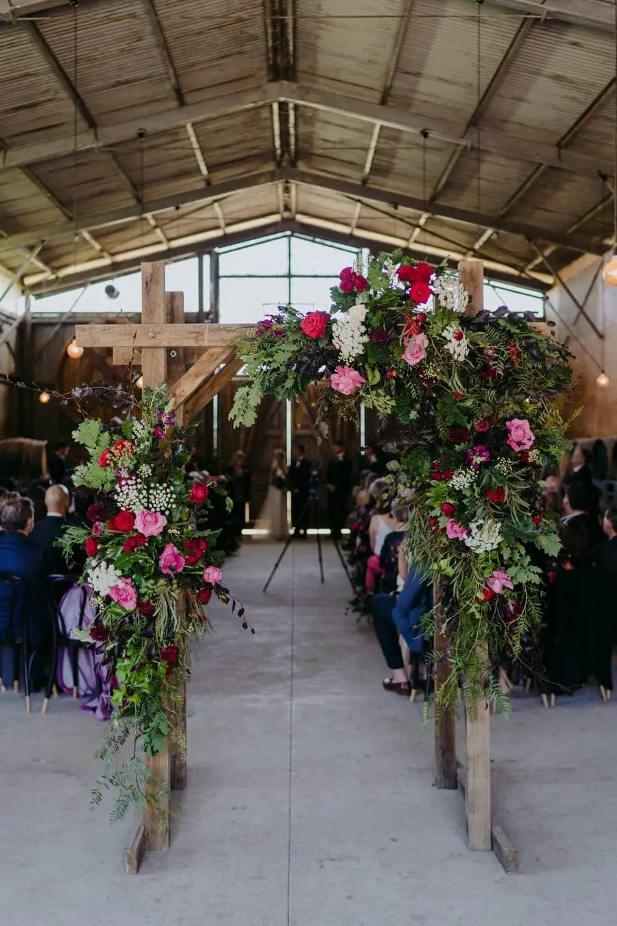 Timber wedding arch decorated with pink flowers and greenery inside a barn