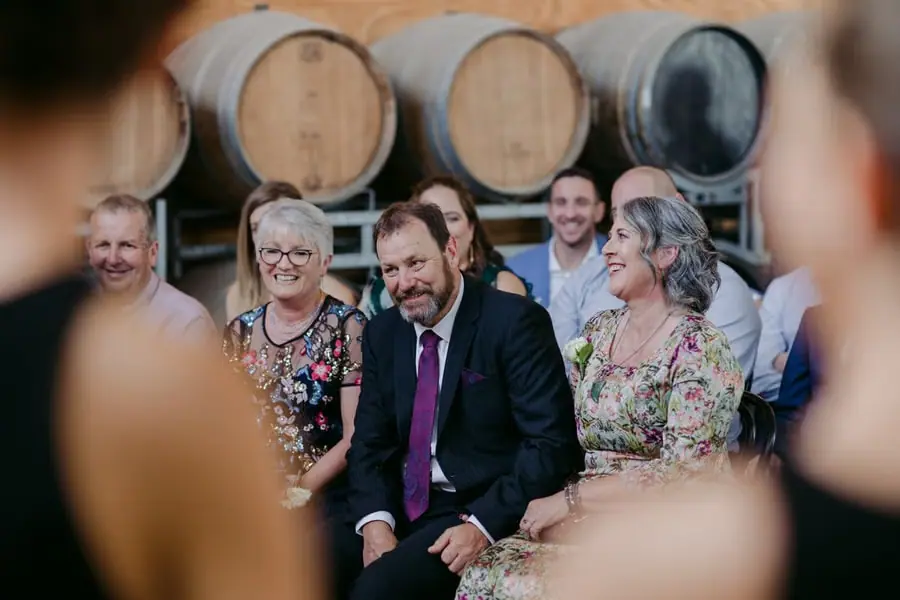Seated wedding guests smiling beneath large timber barrels