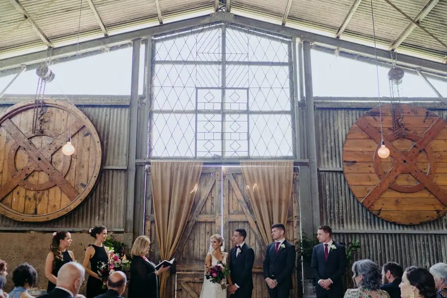 Wide view of a wedding ceremony beneath a high barn roof and large window