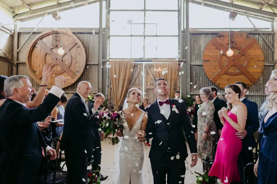 Bride and groom walking through falling petals as guests celebrate inside the barn