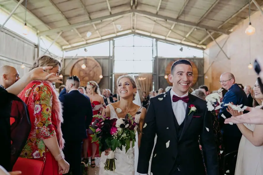 Bride and groom smiling as guests toss petals along the indoor aisle