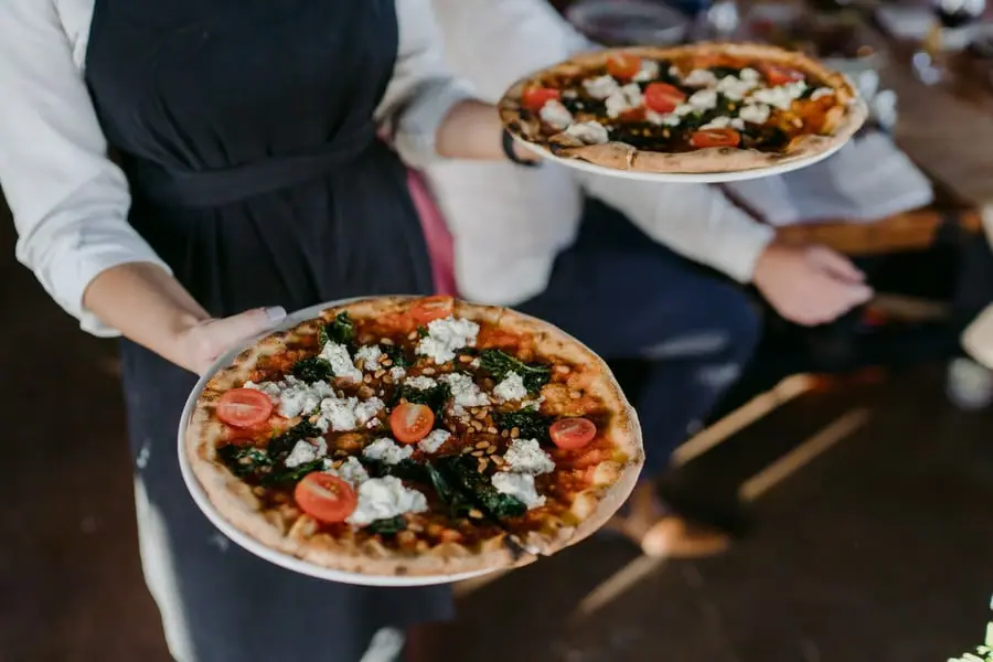 Server carrying two pizzas topped with tomatoes and leafy greens