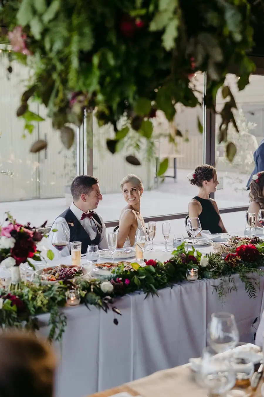Bride and groom seated behind flowers and greenery at the reception table