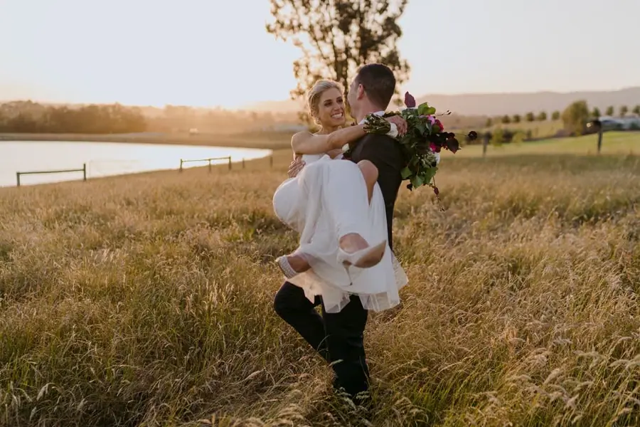 Groom carrying the smiling bride through long grass beside a lake at sunset