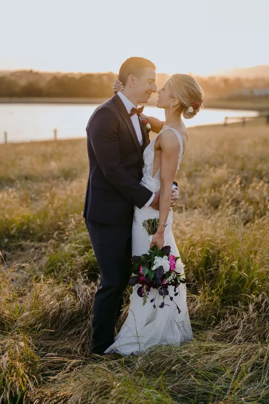 Bride and groom leaning toward each other in a sunlit field beside a lake