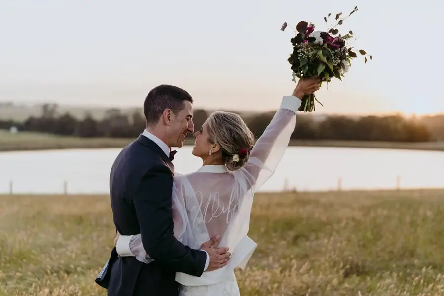 Bride raising her bouquet while the groom embraces her beside a lake