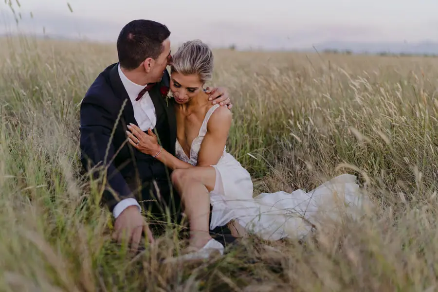 Bride and groom sitting close together in long grass with her dress spread beside them