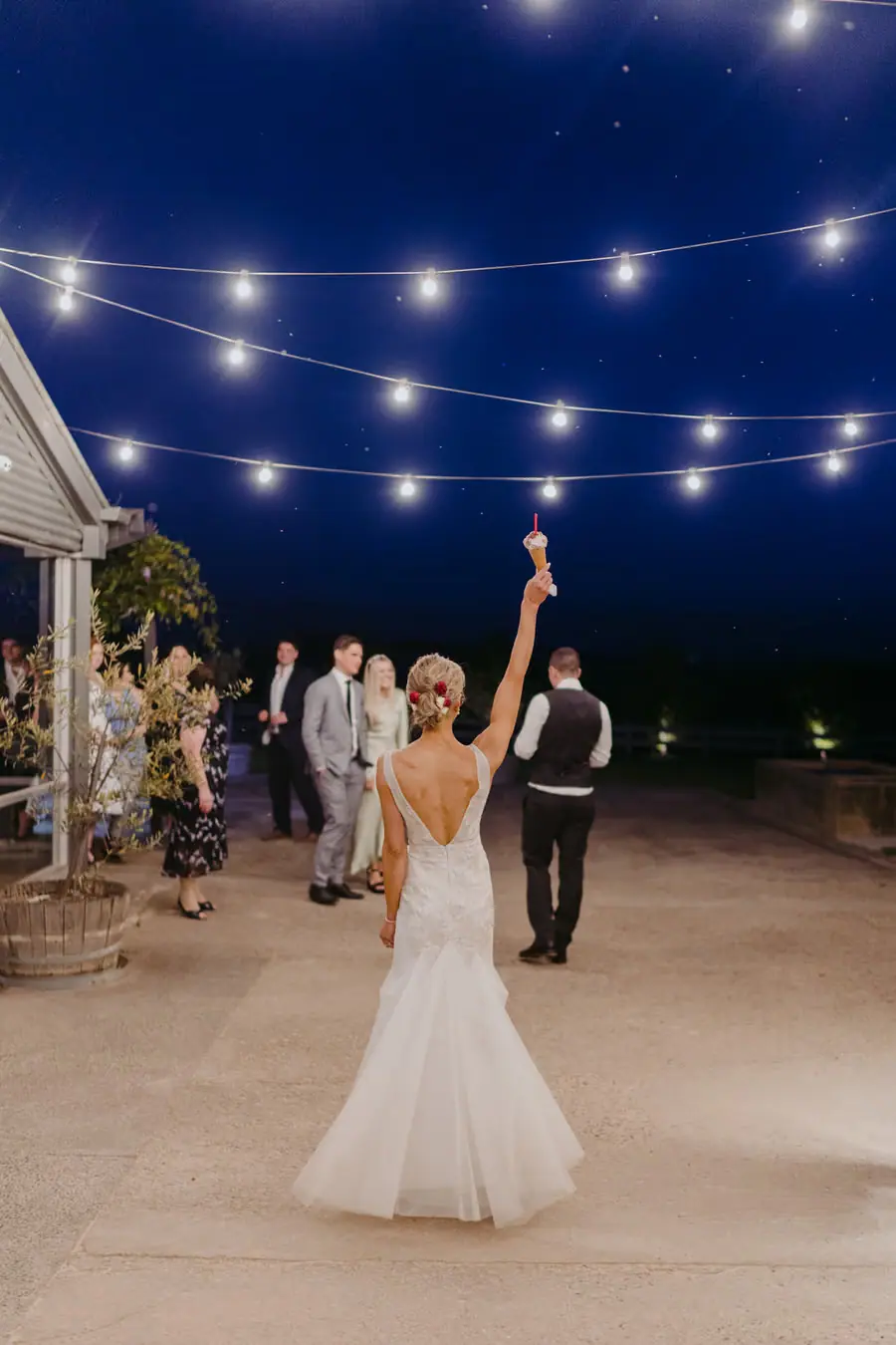 Bride raising an ice cream cone beneath festoon lights in an outdoor courtyard at night