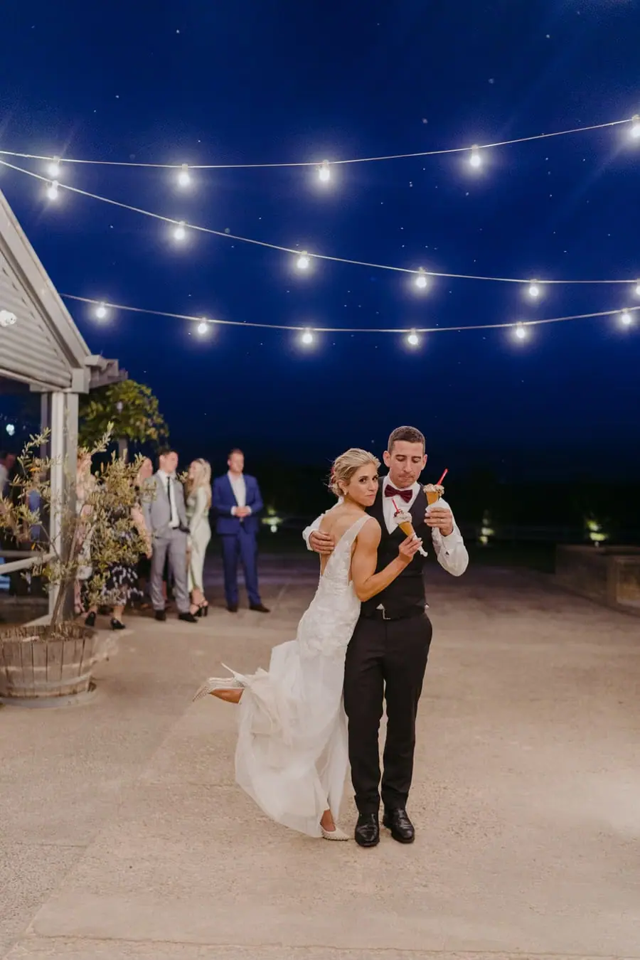 Groom holding the bride from behind beneath outdoor festoon lights
