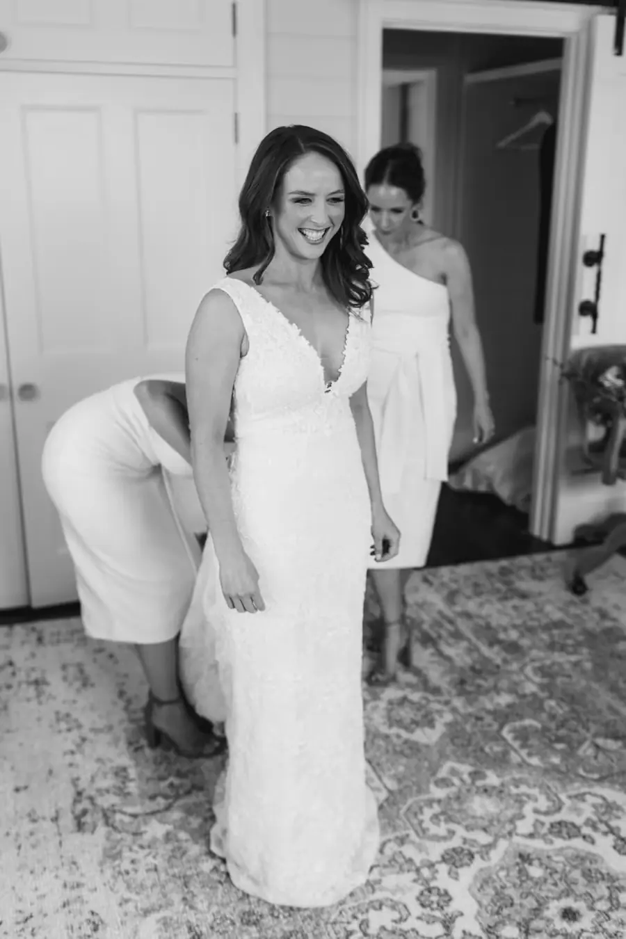 Black-and-white photograph of the smiling bride walking through a bedroom