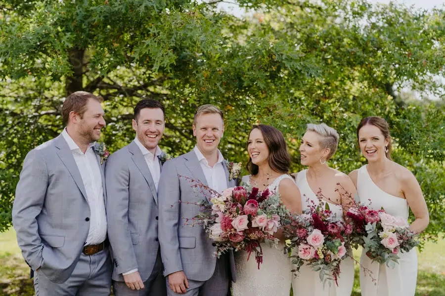 Wedding party laughing together with colourful bouquets beneath trees