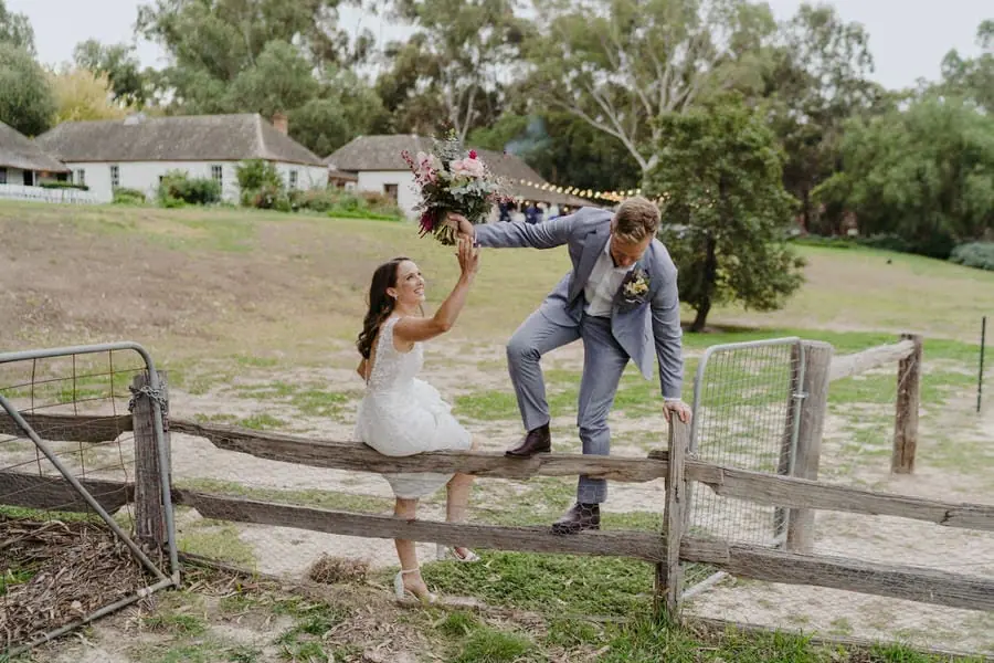 Groom stepping over a timber fence as the bride sits on its top rail