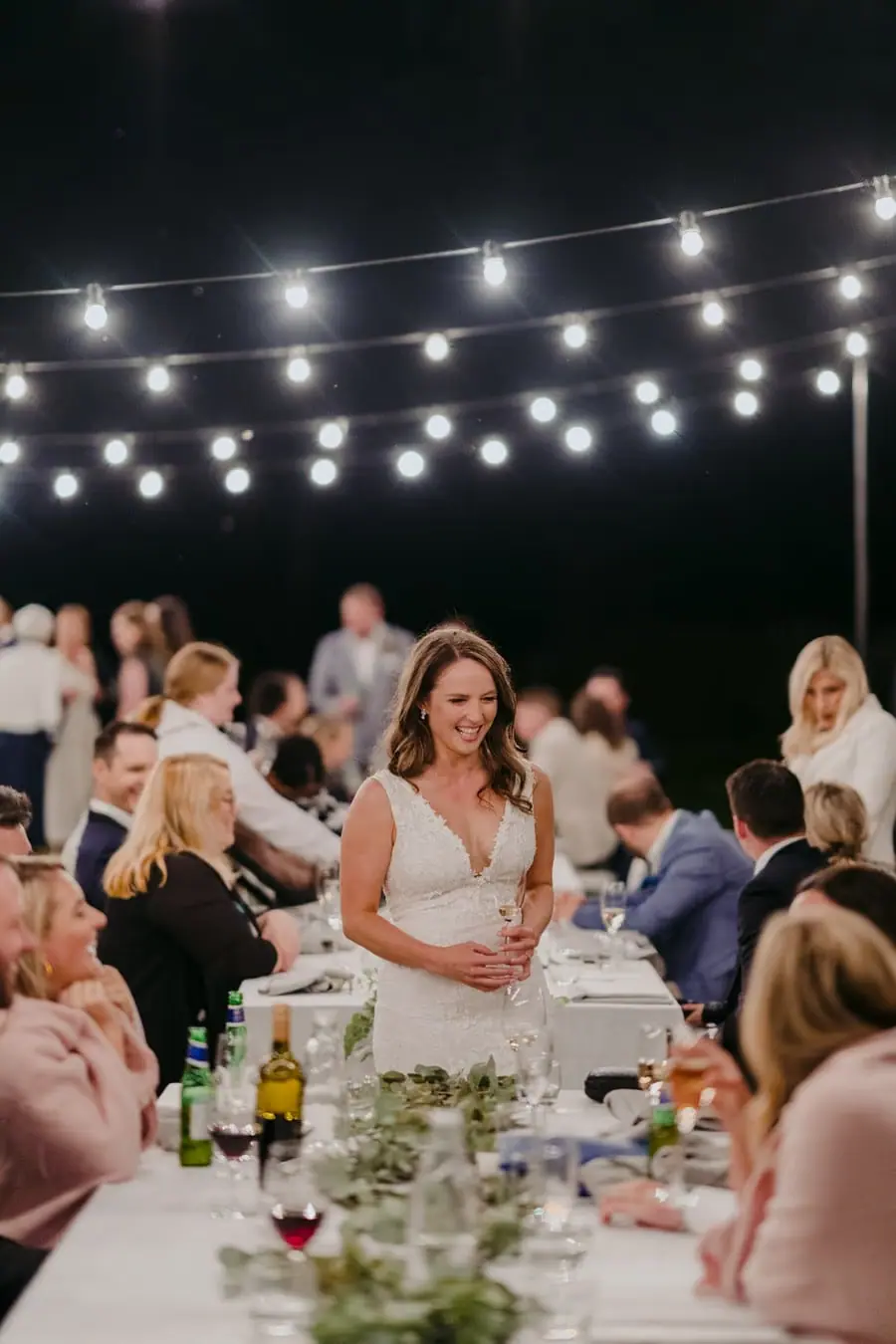 Bride standing among dining guests beneath glowing outdoor lights