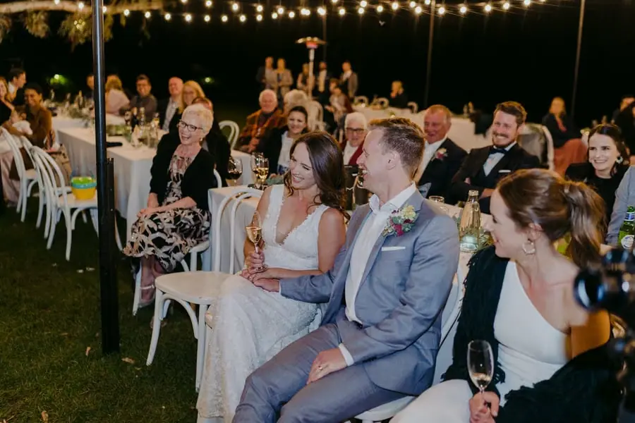 Bride and groom seated among guests beneath warm festoon lights