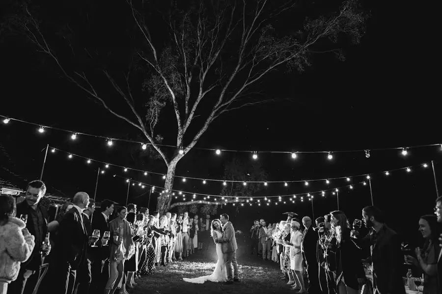 Black-and-white wide view of guests gathered around the dancing couple beneath a tree