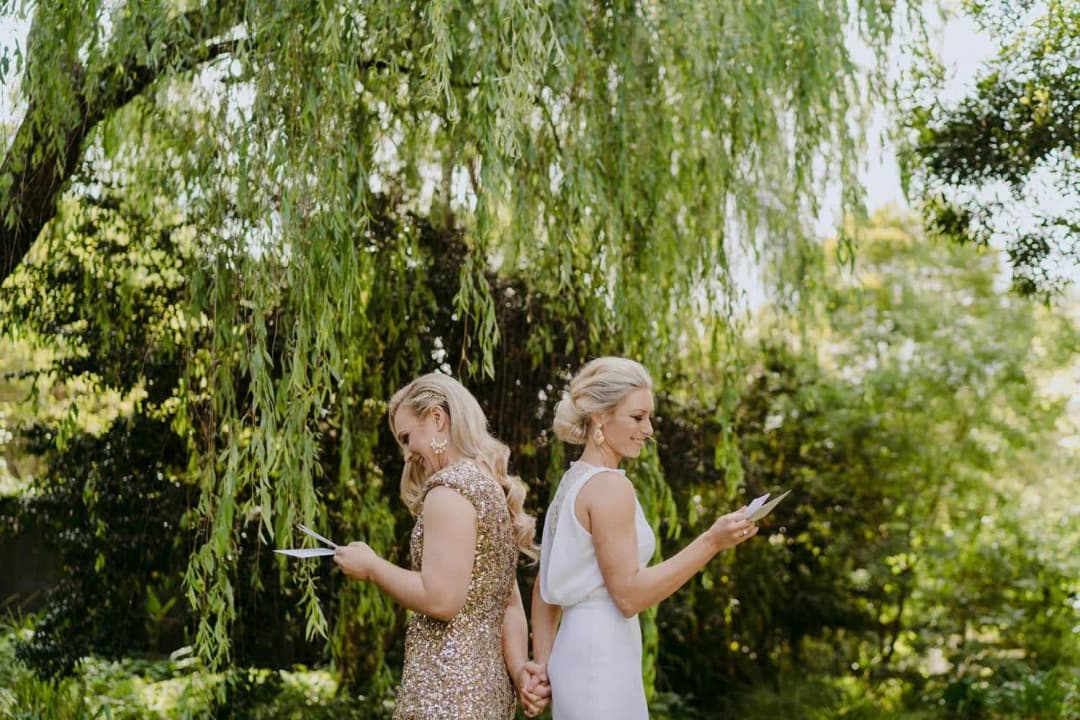 Two brides reading cards while standing back to back beneath a willow tree.