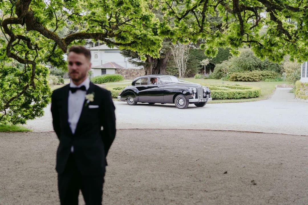Groom waiting beneath leafy branches while a vintage car is parked behind him.