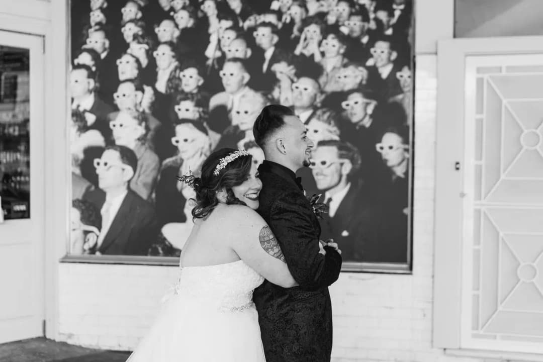 Black-and-white photograph of a bride embracing the groom beneath a large wall painting.