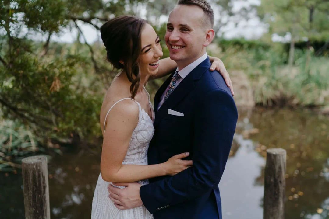 Bride and groom embracing and laughing beside wooden posts in a leafy garden