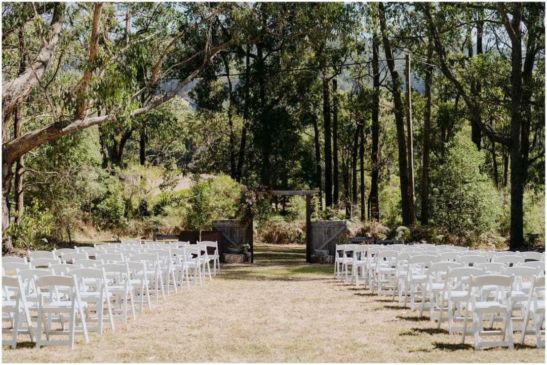 White ceremony chairs facing a rustic wooden arch in a forest clearing.