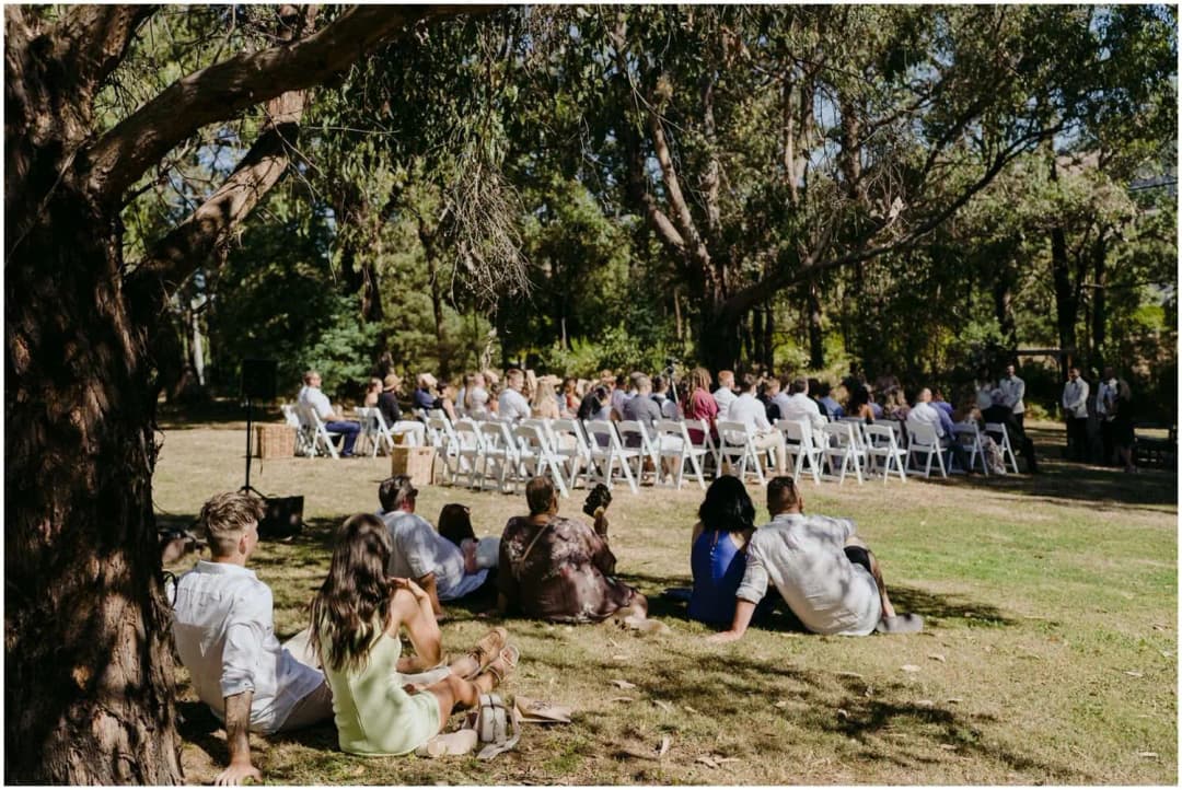 Guests seated on chairs and picnic rugs beneath large leafy trees.