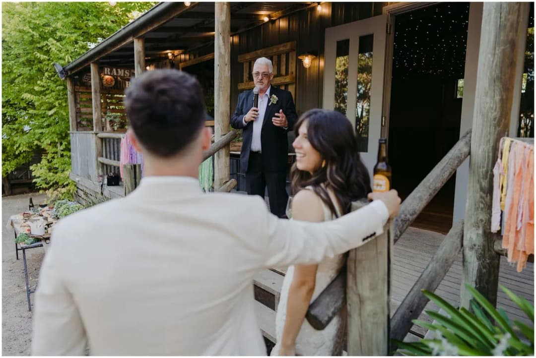 Man speaking into a microphone from a timber veranda as the couple watches.