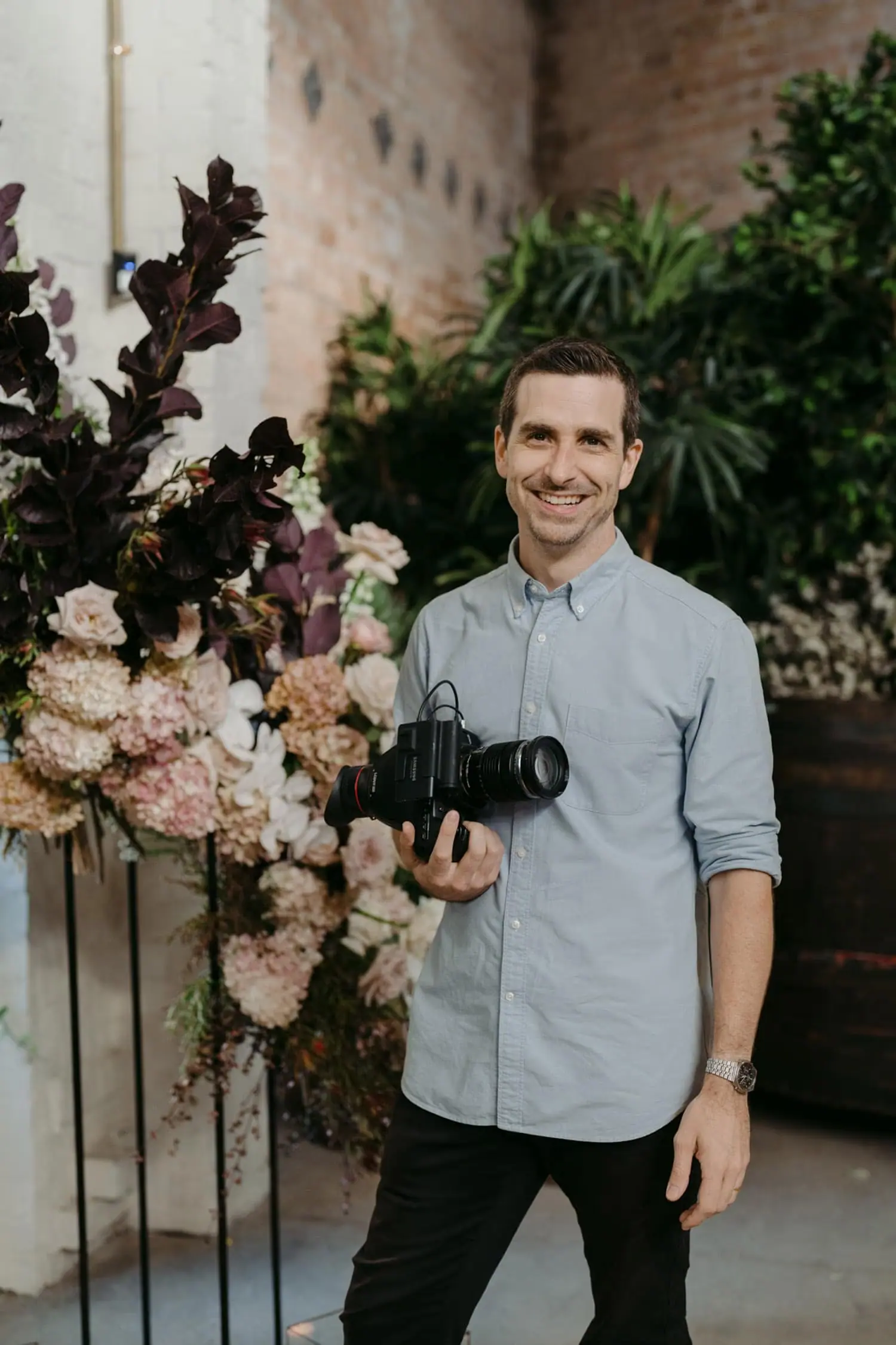 Man holding a camera and smiling beside a floral display.