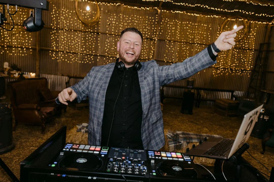 DJ raising an arm behind a mixing console beneath warm string lights.