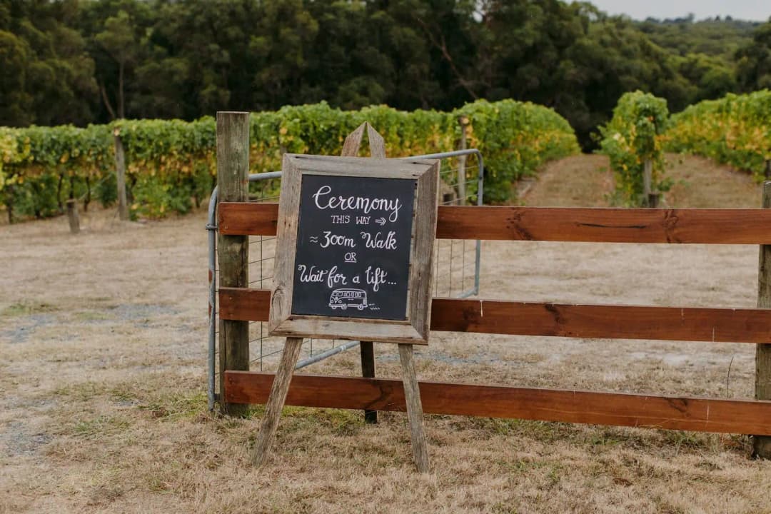 Ceremony chalkboard sign beside a timber gate and vineyard rows.