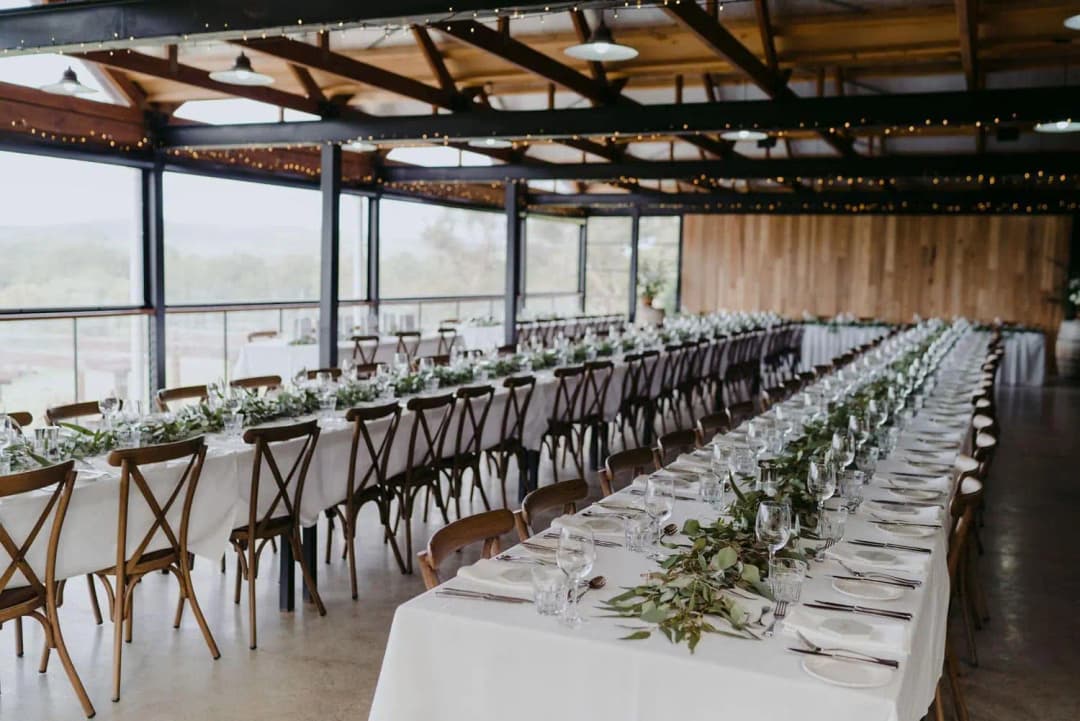 Long white reception tables with greenery beside wide windows and timber roof beams.