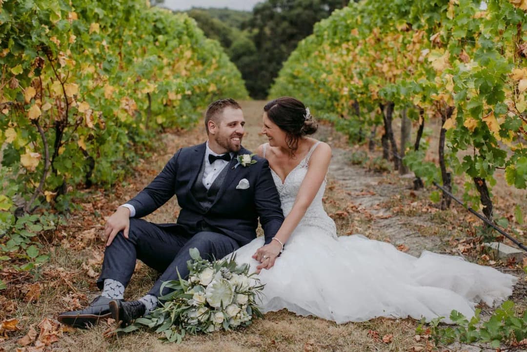 Bride and groom sitting together between leafy vineyard rows.