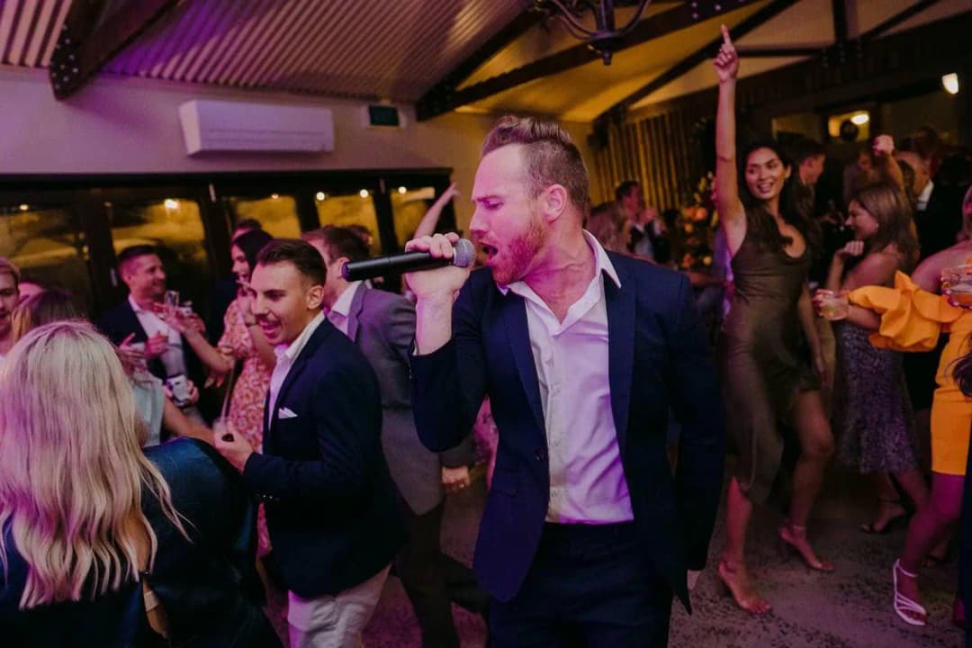 Man singing into a microphone among dancing guests beneath purple lighting.