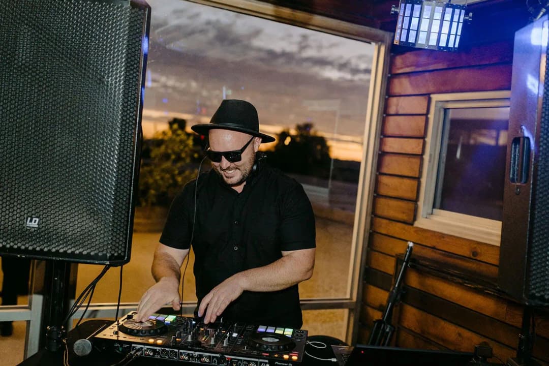 DJ wearing a hat and sunglasses while working at a mixing console.