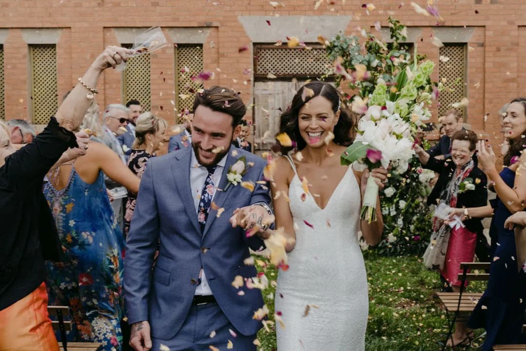 Couple laughing while guests throw colourful confetti outside a brick building.