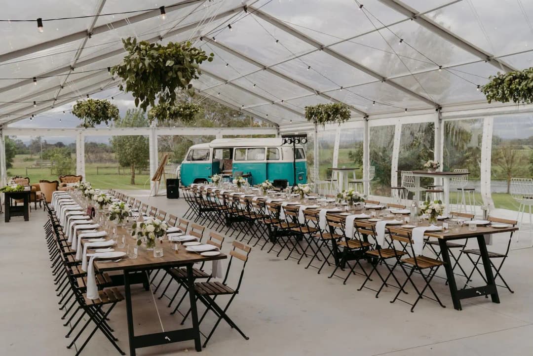 Reception tables beneath hanging greenery inside a clear marquee beside a turquoise vintage van.